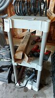 White resin three shelf unit with three shelves, containing coils, cordless drills and boxed items on and around it.