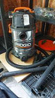 View of Ridgid shop vacuum on shelf with attached hose and various black plastic attachments displayed