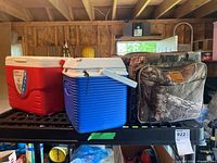 Shows two hard-sided coolers, red/white and blue/white, and one camouflage soft-sided cooler bag on a black shelf in a garage.