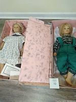 Photo showing both Annette Himstedt dolls in pink patterned boxes, girl doll in a floral dress and boy doll in a green outfit. Certificate of authenticity visible.