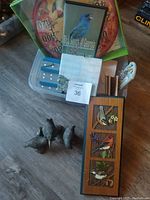 Photo showing boxed colorful hummingbird thermometer, DVD with blue bird cover, wood carved bird picture panel, brass bird figurines, and other small bird-related items on floor