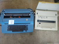 Blue IBM Selectric electric typewriter and beige Brother electric typewriter side by side on floor, showing overall condition and size.