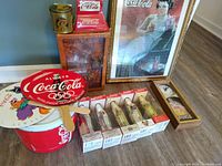 Overview of several Coca-Cola collectibles including limited edition bottles, glasses in wooden case, neon sign, clock, fan, cooler bucket and tin can container.