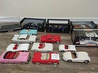 Shows 13 diecast cars arranged on floor with some in display cases and boxes. Various colors including pink, red, white, blue, and black.