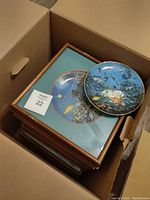 View of a cardboard box containing 8 framed collectors plates stacked with one unframed plate on top, showing the decorated face with marine wildlife scene and wooden frames with wear.