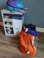 Photo of 6-gallon Igloo beverage cooler in box, purple and blue helmet, blue water bottle, and orange bag with camping equipment on floor.