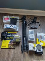 Full view of the lot items including iPhone 7 box, tripod, wireless phone, camera accessories, and binoculars arranged on floor.