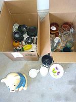 Two cardboard boxes filled with assorted kitchenware including bowls, cups, glassware, and decorative items along with ceramic bear cookie jar and mugs on the floor.
