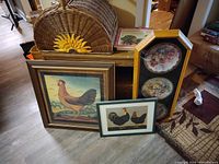 Overview of large wicker picnic basket with sunflower decoration, three pieces of rooster art in frames, and a decorative framed plate set leaning against a piece of furniture.