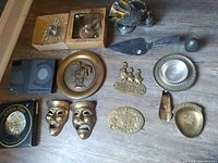 Photo showing full assortment of vintage brass and silver items on wooden floor, including boxed chokin plate, theatrical masks, change holder, belt buckles, and small decorative plates.
