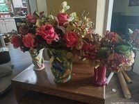 Three decorative vases on a wooden table filled with faux flowers including roses, orchids, and hydrangeas.