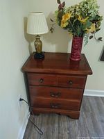 Overall view of wooden nightstand with lamp and vase on top, placed against wall corner.