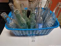 Photo showing 17 vintage soda bottles placed inside a blue plastic basket, various clear and green glass bottles are visible with embossed and printed details.