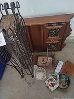 Photo showing the tall black metal CD rack beside the vintage wooden spice rack, and several rooster kitchen items on the ground including a ceramic plate, tile, trivet, small wire basket, and a wooden cutting board.