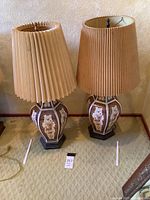 Pair of Asian style ceramic lamps with beige pleated shades on carpeted floor, one shade slightly tilted