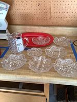 Photo shows one clear cut vintage glass pitcher, six clear cut glass segmented small plates, one small clear glass bowl, and one large red oval serving platter on a wood surface.