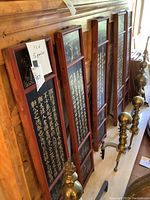 Five tall wooden panels with Asian calligraphy and painted figure scenes leaning against a wall next to brass decorative items.