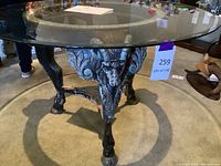 Full view of round glass-top table with ornate goat pan legs and cloven hooves on a carpeted floor.