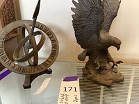 Two sculptures on glass table: iron armillary sphere sundial with Roman numerals and eagle sculpture with wings spread over rock base.