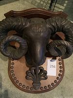 Front view of the metal ram's head sculpture mounted on the wooden plaque showing details of horns, face, and plaque design.