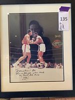 Framed print depicting a boxing match scene, black frame with white mat, signed by Muhammad Ali