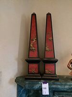 Front view of the pair of small wooden obelisks painted red with gold landscape scenes and black trim.