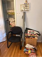Wide shot showing black chair with chenille bedspread draped over the chair, a tall rolled fabric item, and a box stacked with folded quilts and blankets including a floral quilt with pink and yellow patches.