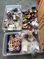 Multiple plastic bins filled with porcelain dolls, some wrapped in plastic bags, and wooden doll furniture.