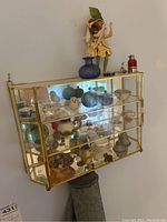 Front and side angle photo of wall-mounted glass and brass display case with assorted figurines and small decorations including ceramic birds and miniature vessels.