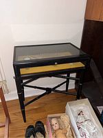 Full view of the black display table showing the glass top and items inside, including fans and cups. Wooden frame with tapered legs and cross support.