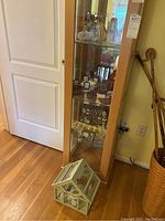 Full view of wooden china cabinet with glass doors, containing multiple items inside and miniature glass garden house placed in front on floor.