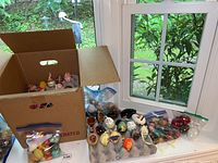 Wide view of multiple decorative eggs including painted wood, glass, and stone types in a cardboard box and laid out on a table