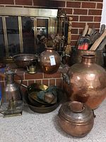 Full lot overview showing fireplace tools behind, copper and brass vessels arranged near hearth: including large copper pot, smaller pots, pans, jug, and brass utensils.