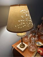Table lamp with brown tapered lampshade featuring floral cutout design and brass base with glass and marble elements, placed on wooden surface surrounded by glass bottles.