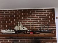 Four handcrafted model ships displayed on a wooden shelf against a brick wall. Includes a white riverboat, two sailing ships, and a paddle wheeler steamboat.