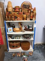 Full view of the white shelving unit with all the items displayed, including nesting dolls, wooden boxes, figurines, decorative plates, wooden vases, and a vintage cheese box.