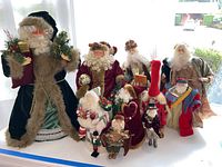 Grouped view of 10 Santa Claus figurines showing varied sizes and decoration styles on white surface in front of window