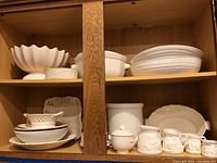 Open wooden cabinet with multiple vintage white ceramic serving items including bowls, platters, and serving dishes arranged on two shelves.
