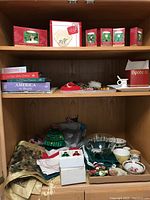 Wide view of shelf with Hallmark Keepsake Peanuts Christmas ornaments in original boxes, assorted Christmas and Americana books, cloth linens, costume jewelry, bowls, and salt and pepper shakers.