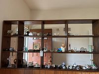 Wooden shelf display showing various miniature tea sets, figurines, plates, and decorative items arranged in cubed compartments in a living room setting.