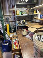 Workshop area with shelves containing a variety of tools, hardware, adhesives, and other equipment including a bucket of materials, multiple hand tools in drawers, and various boxes stored on shelves.