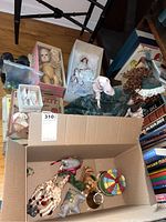Overview of the lot showing various dolls in boxes, a box with assorted vintage toys including a carousel horse and colorful spinning top, and some stacked books and items in the background.