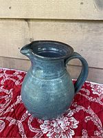 Front and side view of blue glazed pottery jug on red patterned fabric, showing the jug's shape, handle, spout, and glaze details.