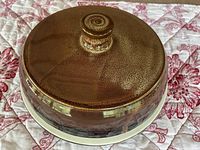Top view of the brown glazed pottery covered dish resting on a patterned fabric, showing the rounded lid with knob handle.