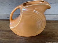 Side view of the orange Fiesta water pitcher showing overall shape, handle, and embossed circular detail.