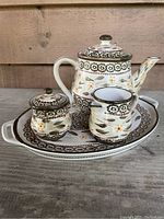 Photo showing the teapot with lid, sugar bowl with lid, creamer, all on top of the oval serving tray with floral and leaf patterns in brown, green, and yellow on cream.