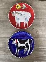 Top view of two ceramic plates, one with sheep on red background with yellow flower, the other with black and white cow on blue background.