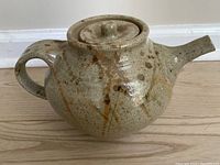 Front angle view of stoneware teapot showing the rounded body, wide handle, and short spout with mottled glaze decoration.
