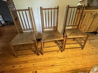 Set of three antique wooden chairs with straight backs and woven seats shown side by side on wooden floor.