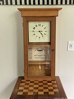 Full view of the oak wall clock showing its height, crown top, glass door and white clock face with black numerals.